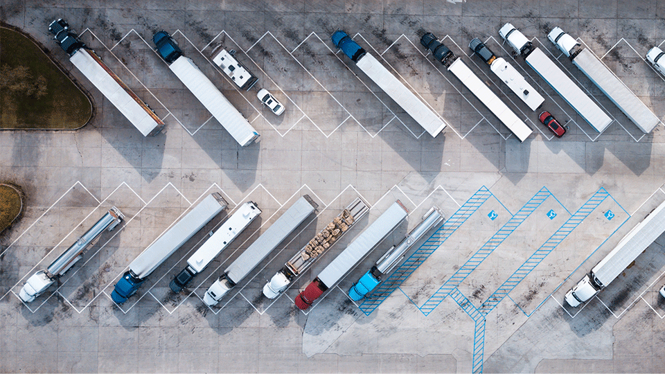 Many semi trucks with a variety of trailers parked at a truck stop.